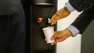 Man in business attire pouring water into a white cup from a dispenser.