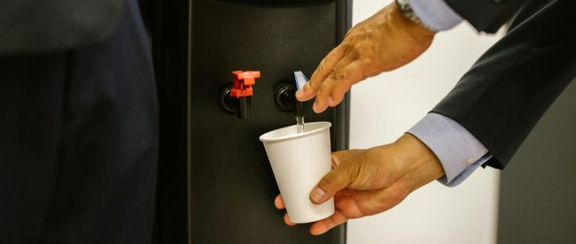 Man in business attire pouring water into a white cup from a dispenser.